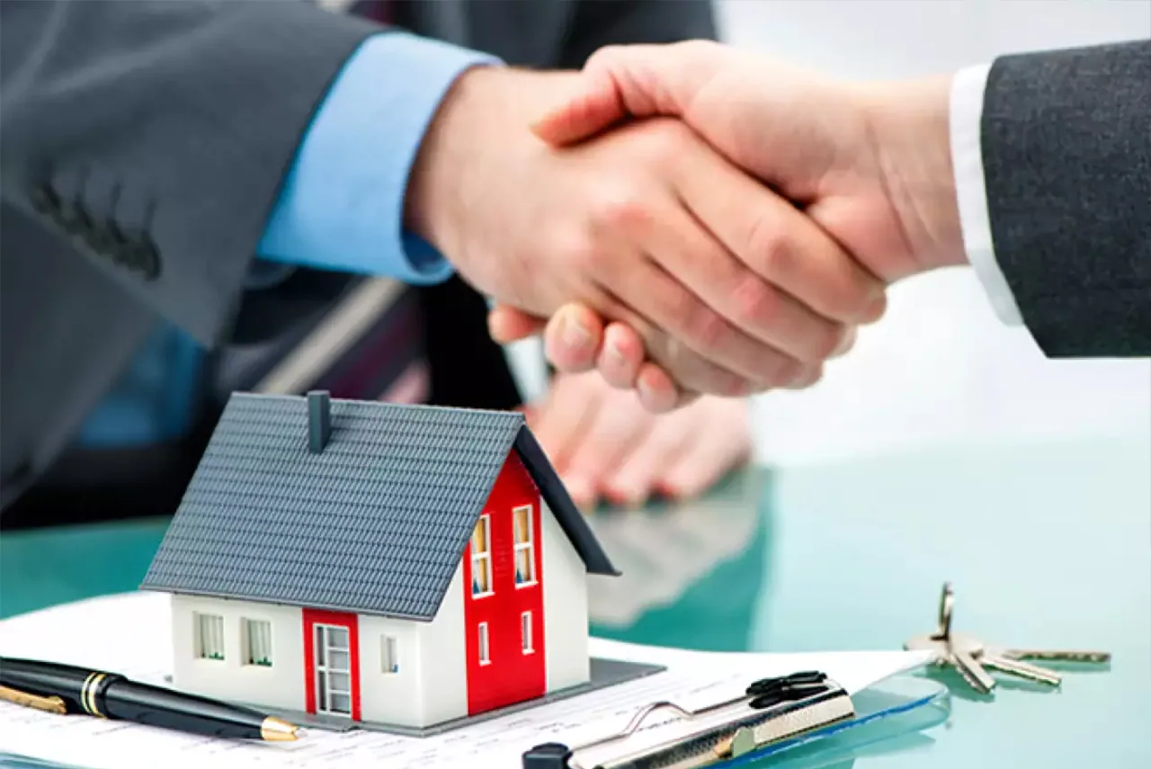 A handshake above a model house, with keys and a pen on the desk.