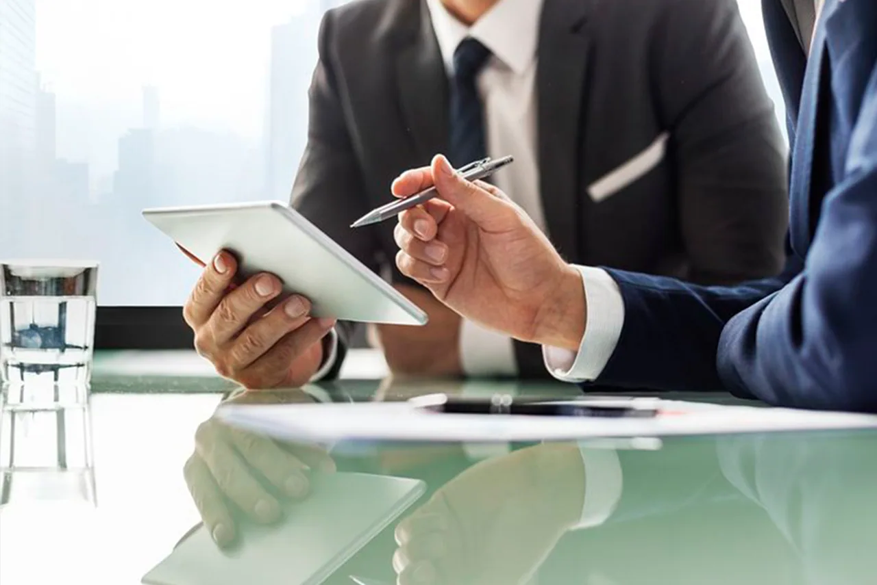 Two professionals review a document on a tablet across a glass table.