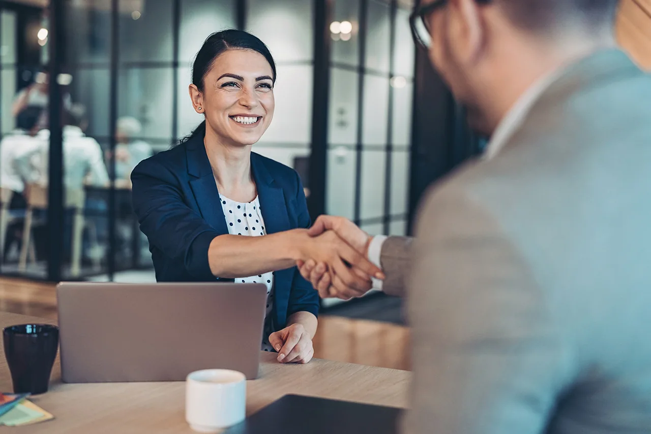 A smiling woman shakes hands with a client across a desk with a laptop.