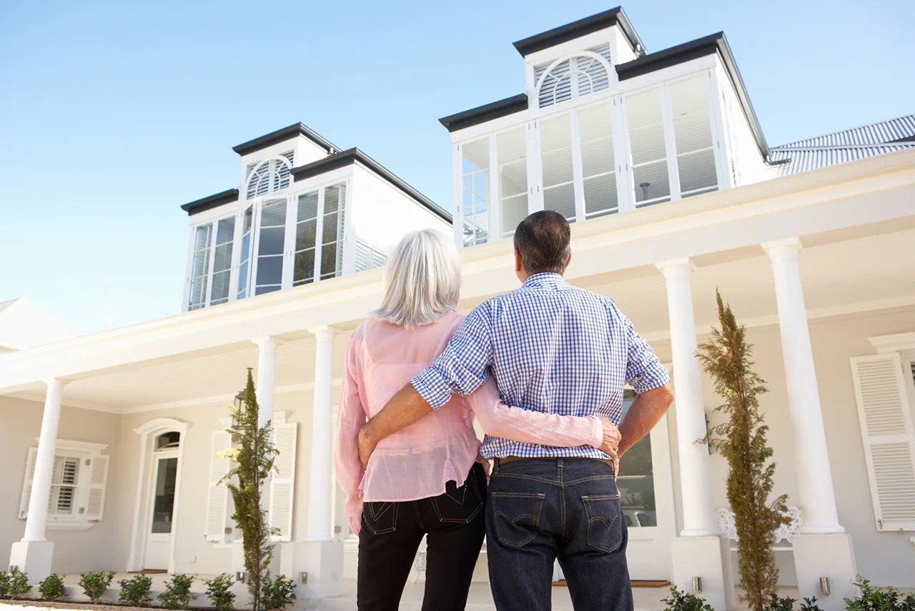 An older couple, arm in arm, looks up at a white two-story house.