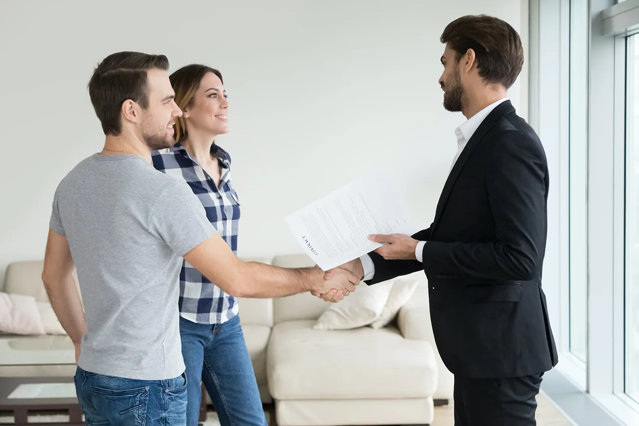 A couple shakes hands with an agent holding a contract in a bright living room.